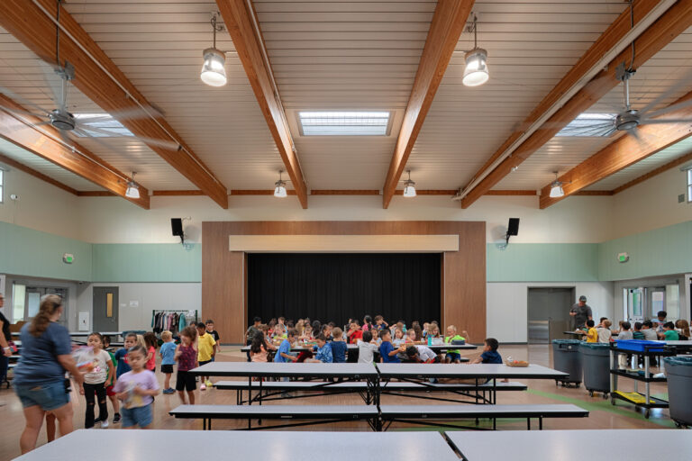 Children eating and walking in a bright school cafeteria with a small stage, wood-beam ceiling, and long tables