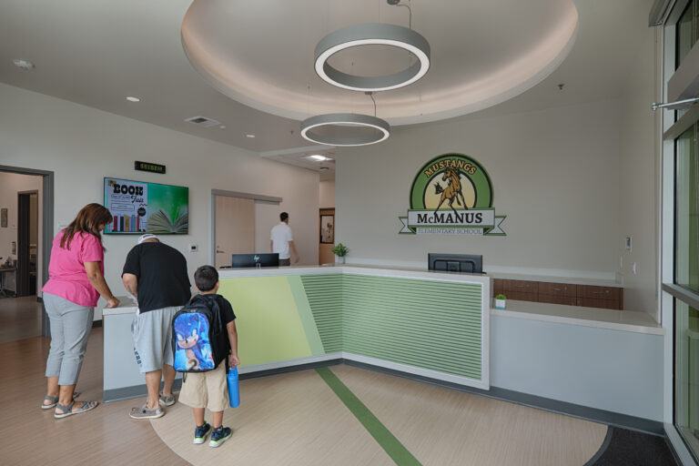 School reception area with families at the front desk and a McManus Elementary School Mustangs logo on the wall