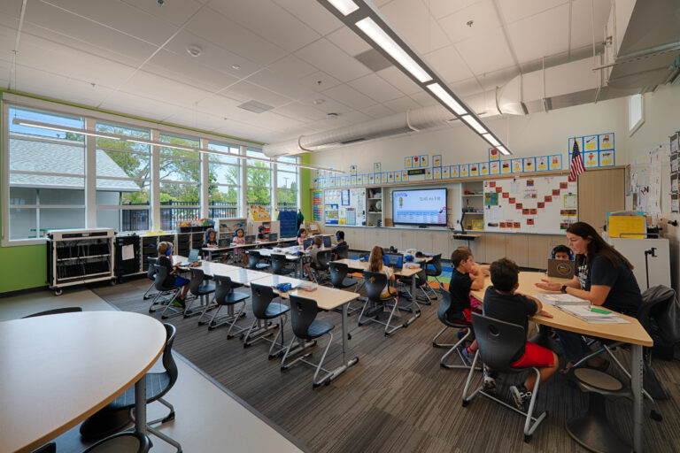 Bright elementary classroom with large windows, students using laptops at desks, and a teacher helping a small group.