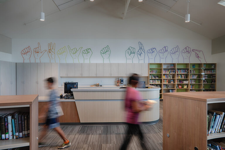 Children move through a library with bookshelves and a colorful hand-sign mural above the circulation desk.