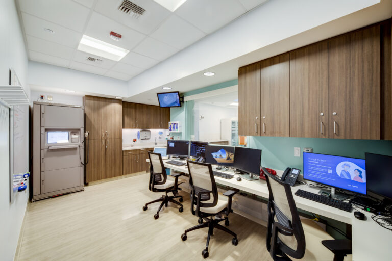 Modern hospital nurses’ station with computers, rolling chairs, wood cabinets, and a medication dispensing unit