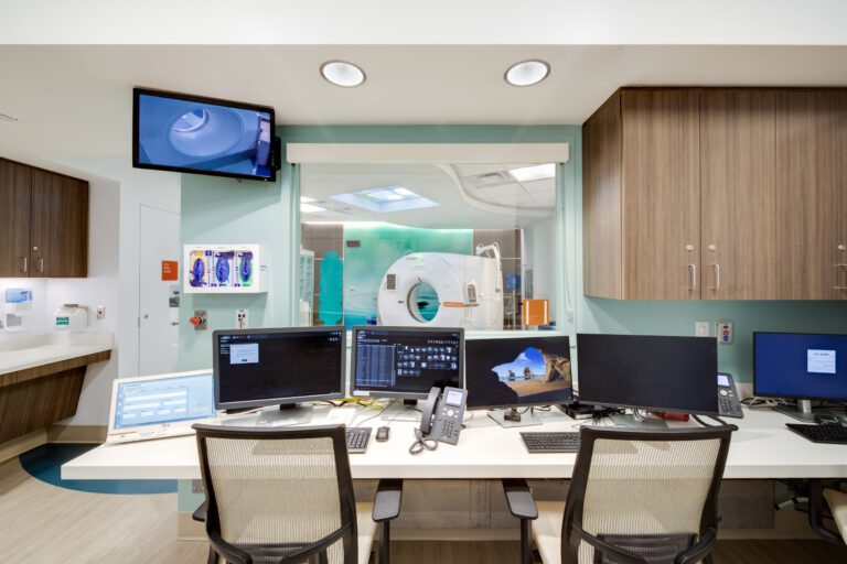 Hospital imaging control room with computer monitors and chairs, overlooking a CT scanner through a large window