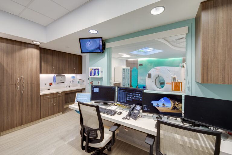 Hospital imaging control room with computer workstations and a CT scanner visible through a large observation window