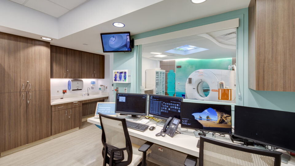 Hospital imaging control room with computer workstations and a CT scanner visible through a large observation window