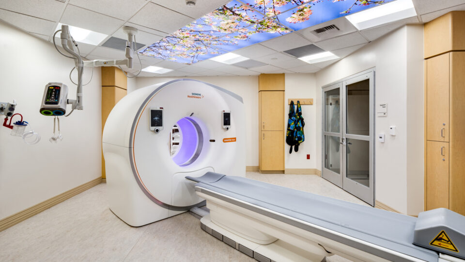 Modern CT scan room with scanner bed, wall-mounted controls, and a ceiling mural of blooming tree branches.