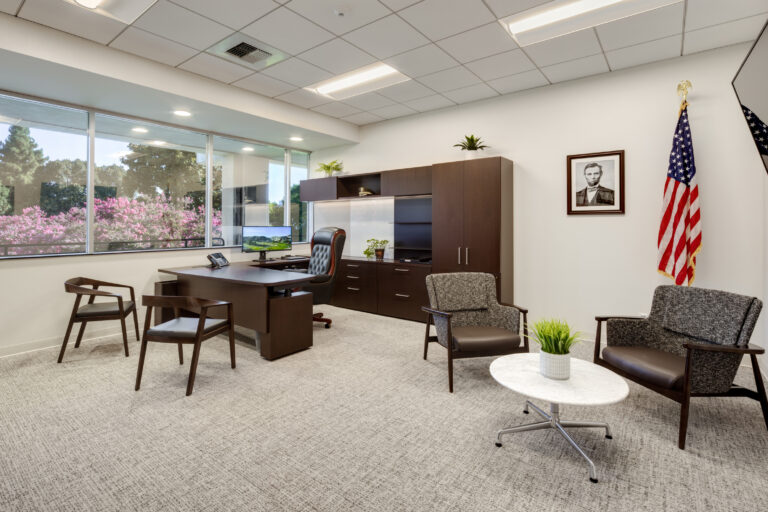Modern office with desk, guest seating, large windows, an American flag, and a framed portrait on the wall.