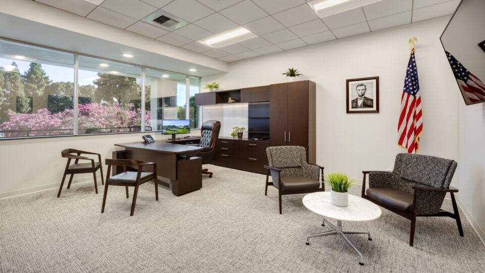 Modern office with desk, guest seating, large windows, an American flag, and a framed portrait on the wall.
