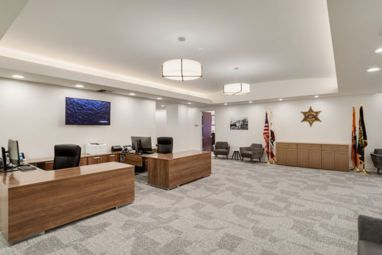 Modern office lobby with wooden desks, gray chairs, wall-mounted TV, flags, and a sheriff’s star emblem on the wall