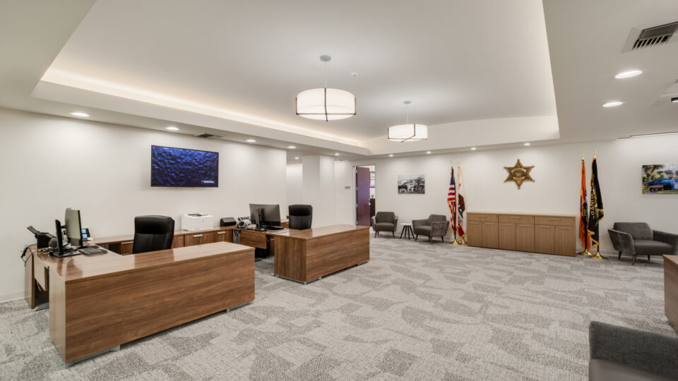 Modern office lobby with wooden desks, gray chairs, wall-mounted TV, flags, and a sheriff’s star emblem on the wall