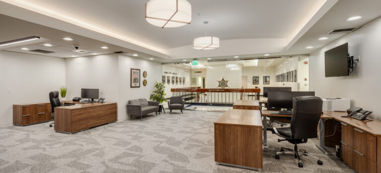 Modern office suite with wooden desks, computer monitors, gray chairs, and a glass wall overlooking a hallway.
