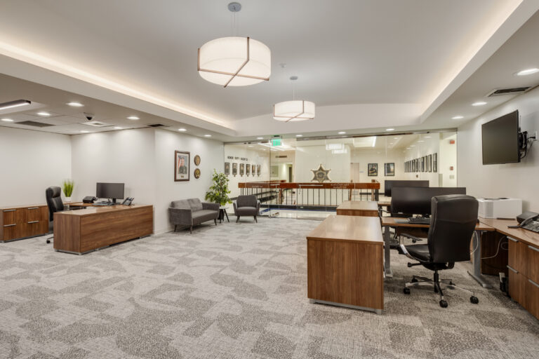 Modern office suite with wooden desks, computer monitors, gray chairs, and a glass wall overlooking a hallway.