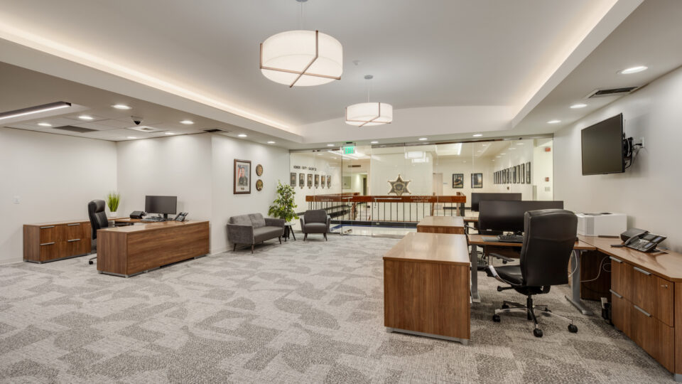 Modern office suite with wooden desks, computer monitors, gray chairs, and a glass wall overlooking a hallway.