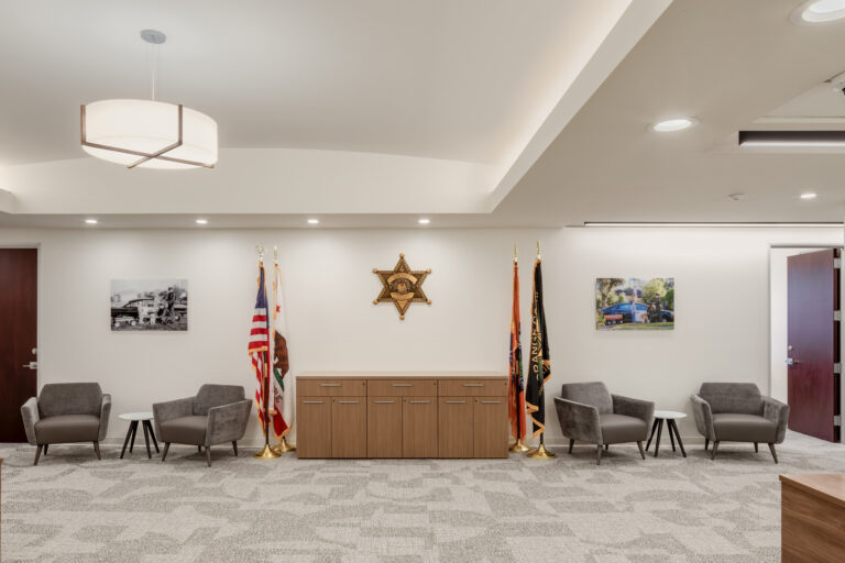 Modern sheriff’s office lobby with gray armchairs, flags, a central cabinet, and a sheriff star emblem on the wall