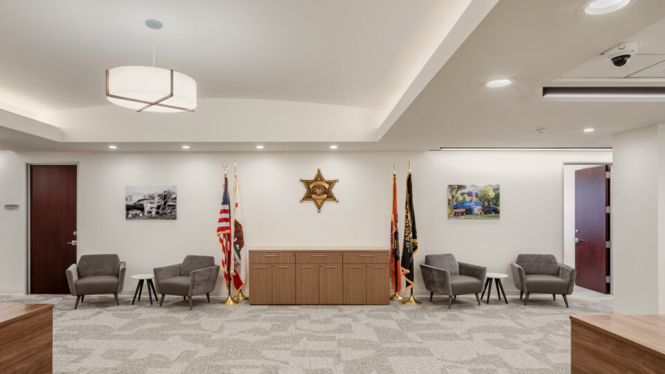 Modern sheriff’s office lobby with gray armchairs, flags, a central cabinet, and a sheriff star emblem on the wall