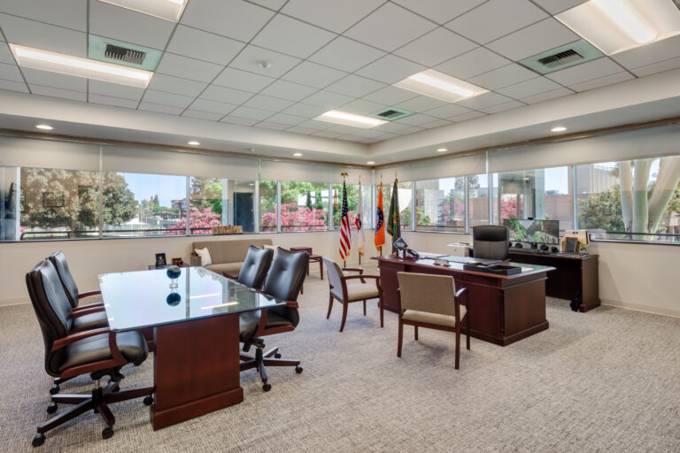 Spacious executive office with desk, meeting table, flags, and large corner windows overlooking trees and buildings