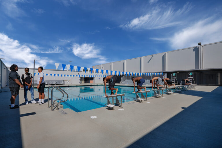 Swimmers crouch on starting blocks beside an outdoor pool while three people stand talking under a bright blue sky.
