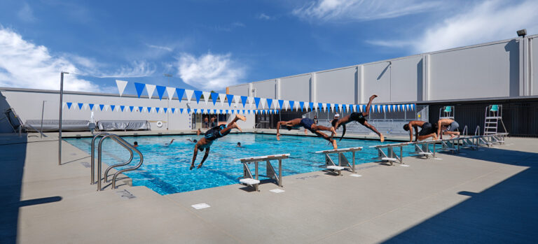 Swimmers dive from starting blocks into an outdoor lap pool beneath blue-and-white pennant flags.