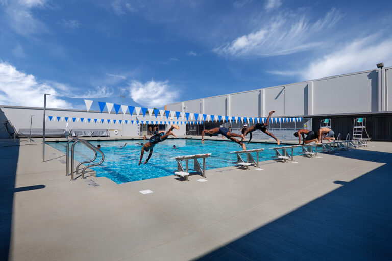 Swimmers dive from starting blocks into an outdoor lap pool beneath blue-and-white pennant flags.