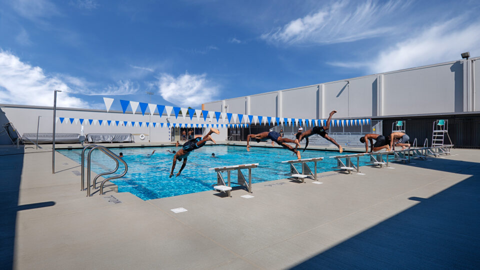 Swimmers dive from starting blocks into an outdoor lap pool beneath blue-and-white pennant flags.