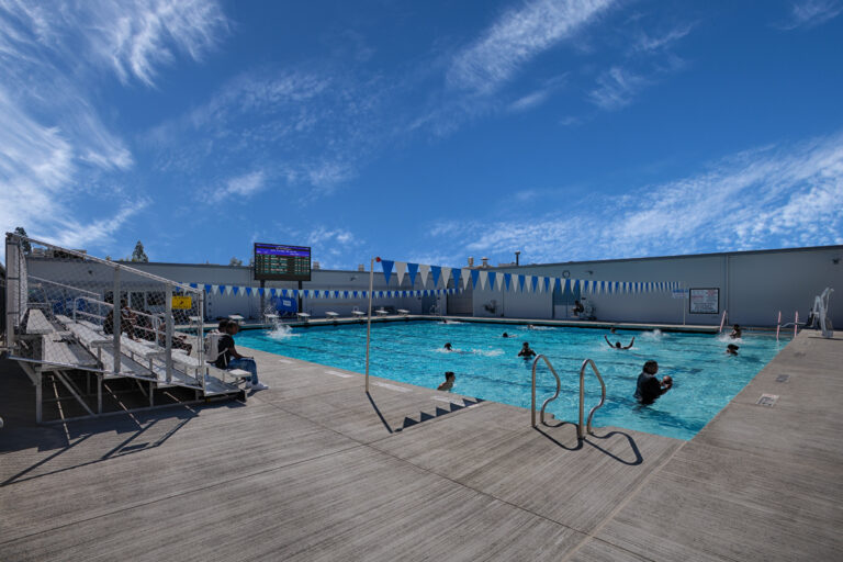 Outdoor swimming pool with several people in the water, bleachers at left, and blue pennant flags under a wide sky