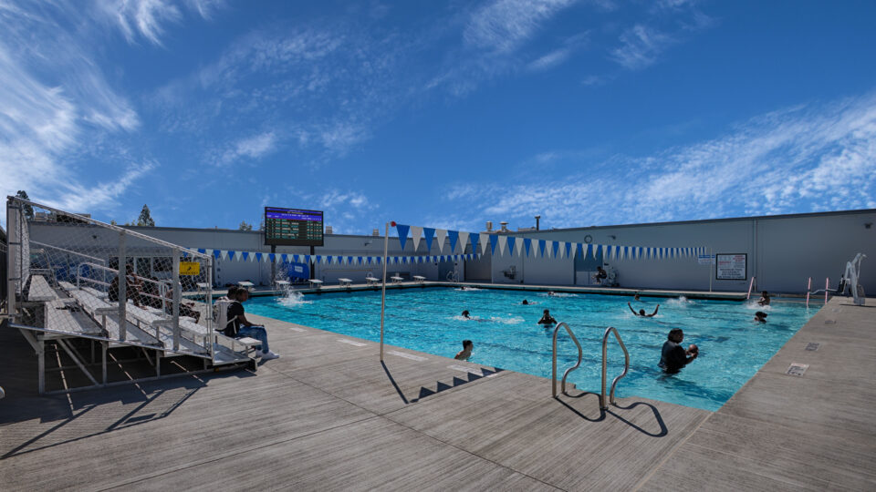 Outdoor swimming pool with several people in the water, bleachers at left, and blue pennant flags under a wide sky