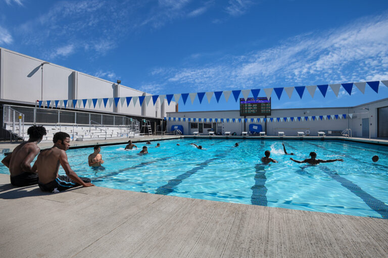 Students swimming and playing in an outdoor pool beneath blue-and-white lane flags on a sunny day.
