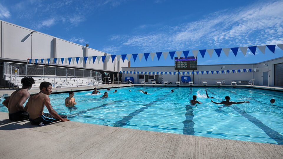 Students swimming and playing in an outdoor pool beneath blue-and-white lane flags on a sunny day.