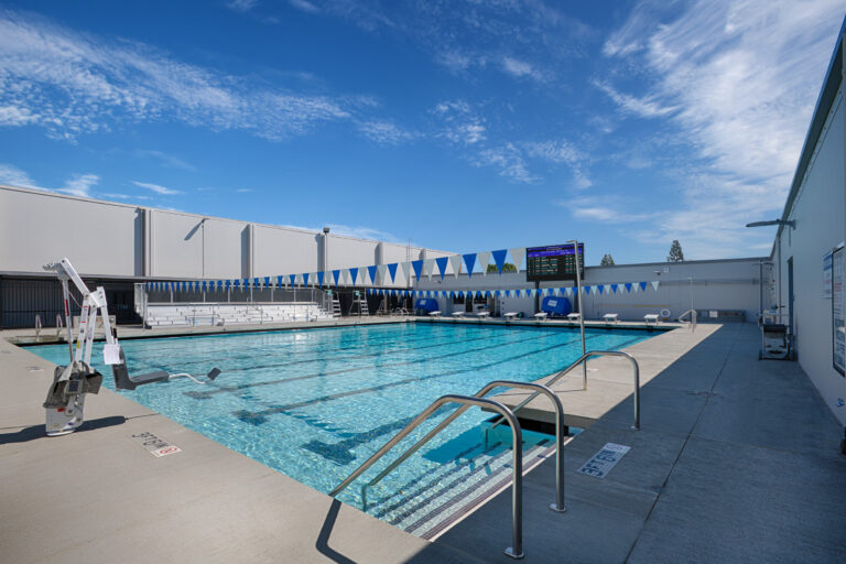 Outdoor lap pool with lane markings, starting blocks, bleachers, and a pool lift under a bright blue sky.