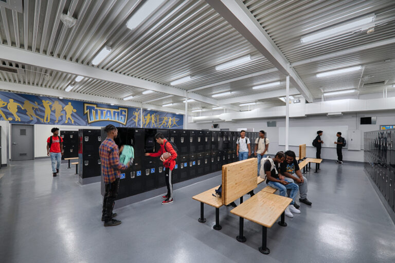 Students in a school locker room with black lockers, wooden benches, and a Titans sports mural on the wall