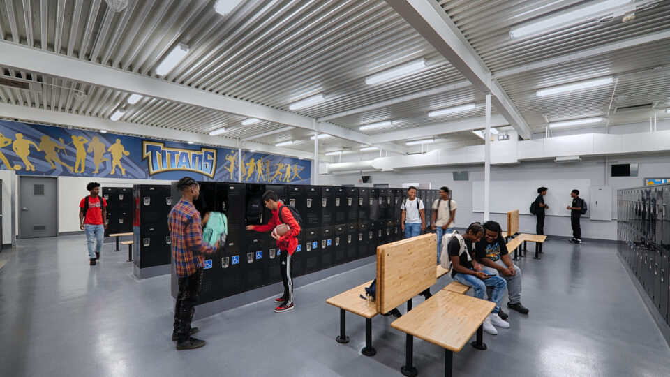 Students in a school locker room with black lockers, wooden benches, and a Titans sports mural on the wall