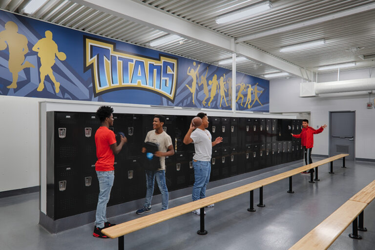 Students in a locker room beneath a Titans sports mural, with black lockers, wooden benches, and one student holding a football