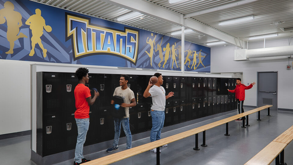 Students in a locker room beneath a Titans sports mural, with black lockers, wooden benches, and one student holding a football
