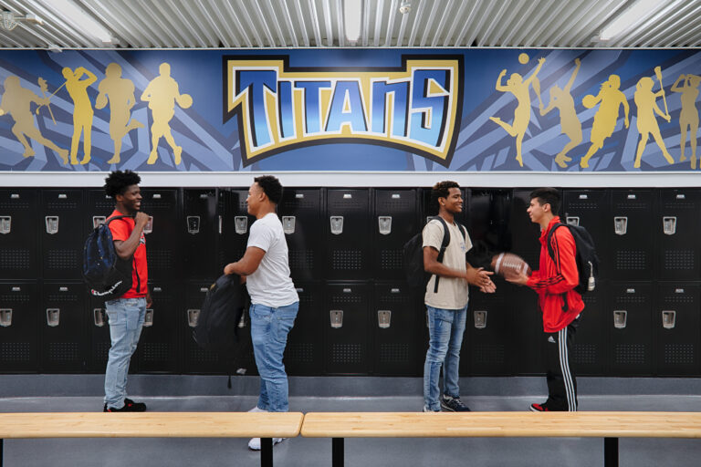 Four students with backpacks chat in a locker room beneath a large Titans mural and rows of black lockers.