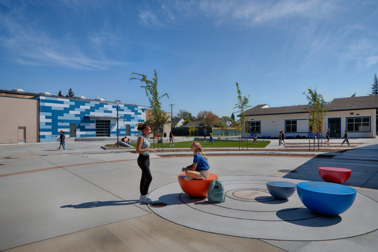 Students gather in a sunny school courtyard with modern buildings, trees, and colorful round seats.