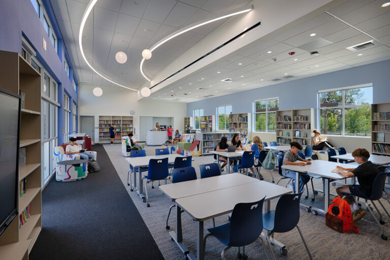 Students reading and working at tables in a bright, modern school library with bookshelves and large windows