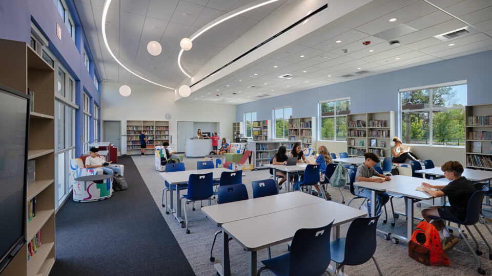 Students reading and working at tables in a bright, modern school library with bookshelves and large windows