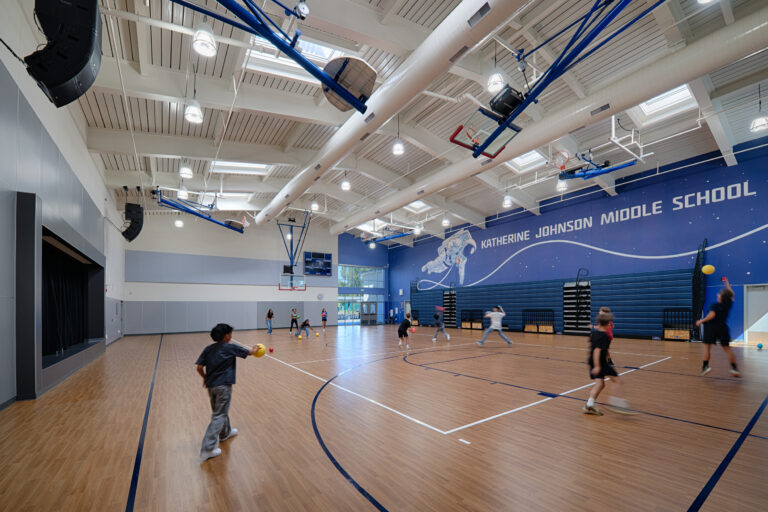 Students play with balls in the Katherine Johnson Middle School gym beneath basketball hoops and a space-themed mural.