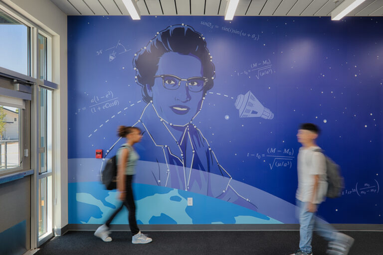 Two blurred students walk past a large blue mural of a woman with glasses, stars, spacecraft, and math equations.