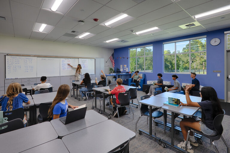 Teacher leading a classroom lesson as students sit at tables, some raising hands, in a bright room with large windows.