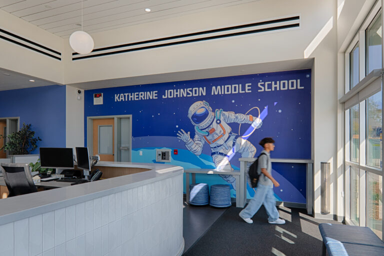 Bright school lobby with reception desk and a mural reading Katherine Johnson Middle School, as a student walks past.