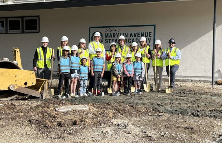 Adults and children in hard hats and safety vests hold shovels at a groundbreaking for Marylin Avenue STEAM Academy.