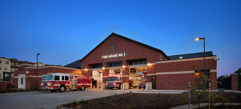 Brick fire station at dusk with illuminated Fire House No. 8 sign and fire engines parked in open bays.