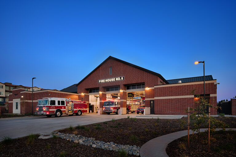 Brick fire station at dusk with illuminated Fire House No. 8 sign and fire engines parked in open bays.