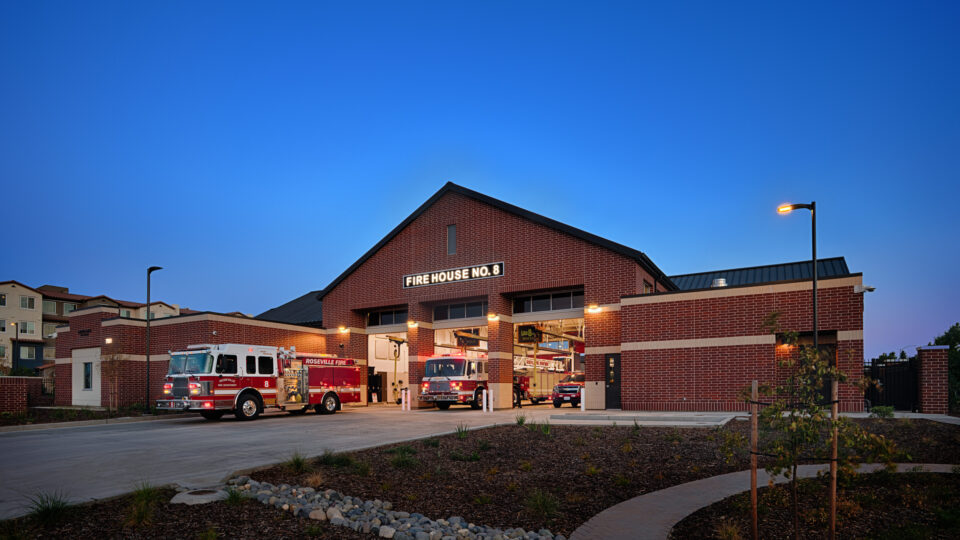 Brick fire station at dusk with illuminated Fire House No. 8 sign and fire engines parked in open bays.