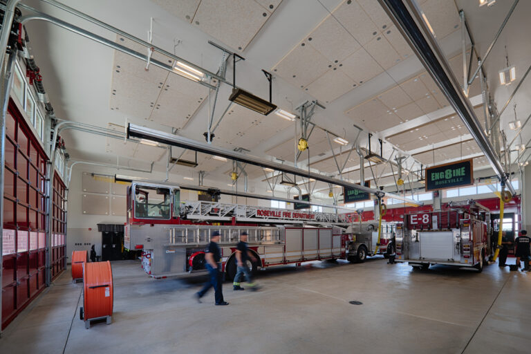 Interior of a modern fire station bay with Roseville Fire Department ladder truck and engine parked inside.