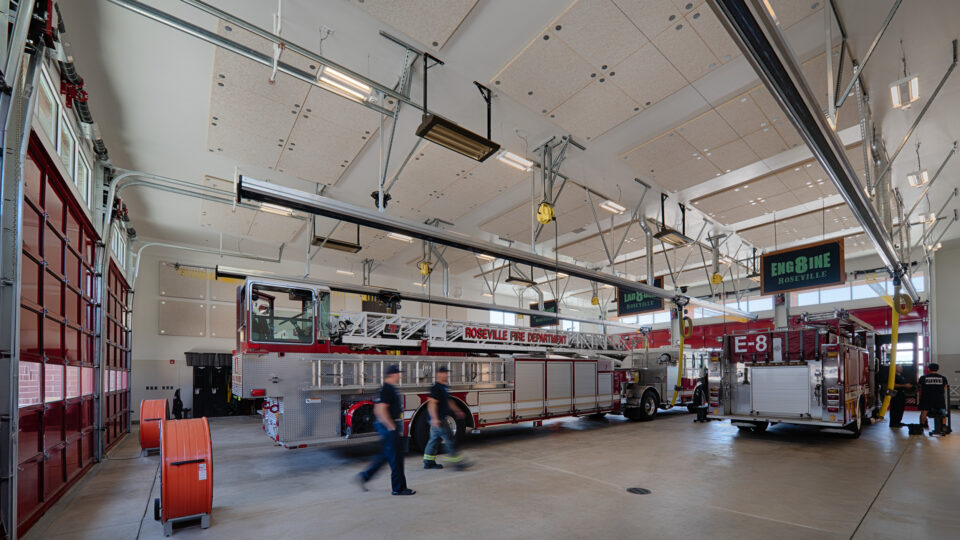 Interior of a modern fire station bay with Roseville Fire Department ladder truck and engine parked inside.