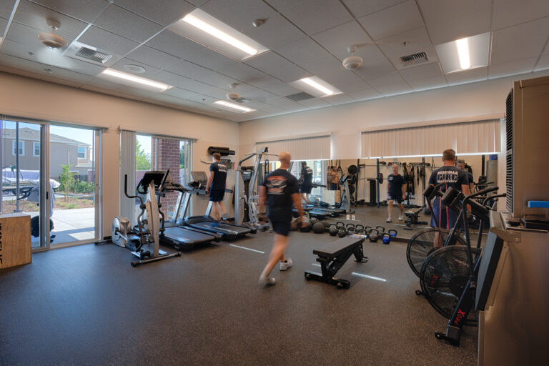 Firefighters exercising in a well-equipped indoor gym with treadmills, kettlebells, benches, and mirrored walls.