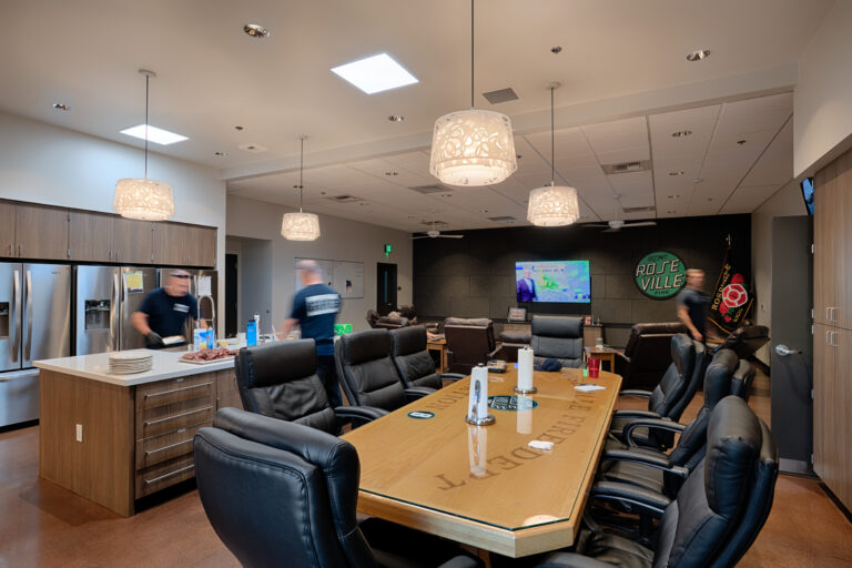 Modern fire station lounge with a long conference table, kitchen island, recliners, and Roseville Fire signage on the wall.