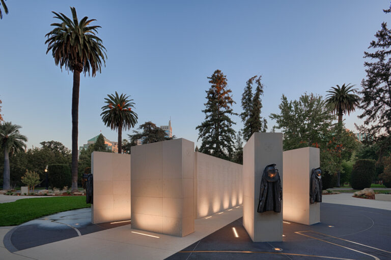 Illuminated stone memorial with hanging firefighter coats in a park at dusk, surrounded by tall palm trees.