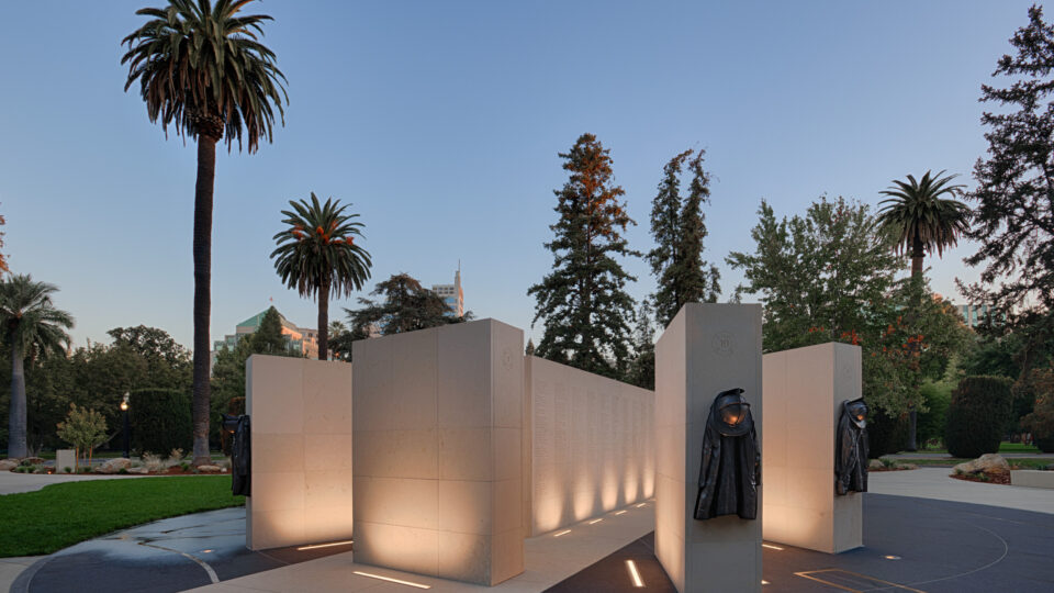 Illuminated stone memorial with hanging firefighter coats in a park at dusk, surrounded by tall palm trees.